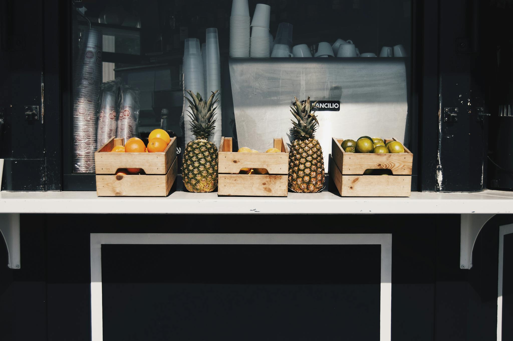 Wooden crates of pineapples, oranges, and other fruits at a market stall on a sunny day.